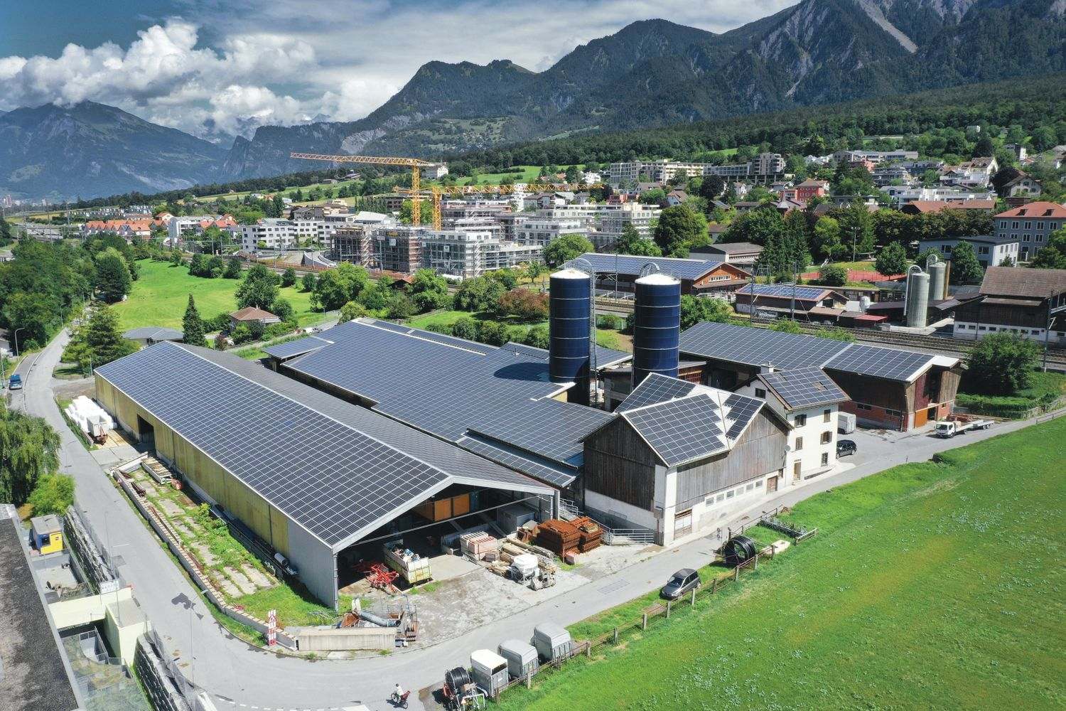Luftbild des Hofs am Reitnauerweg: grosse Stalldächer mit Solaranlage, zwei blaue Silos, dahinter Chur und die Berge